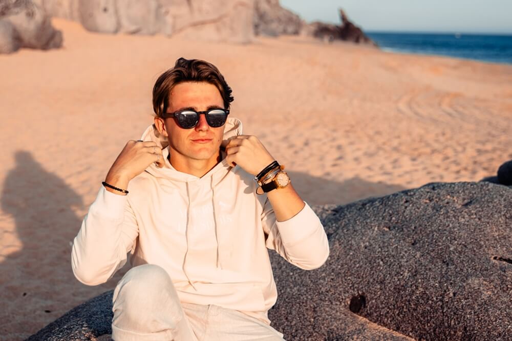 Male model relaxing on a beach, wearing GOWOOD wooden sunglasses. The ocean and sand are visible in the background