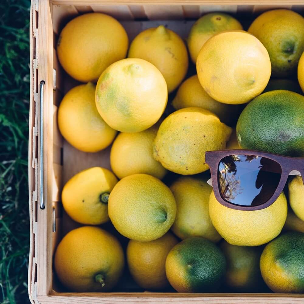 Wooden sunglasses resting on top of a crate filled with bright yellow lemons and a few green limes