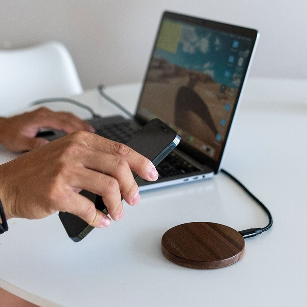 Person's hand placing a smartphone on a wooden wireless charger. A laptop is visible in the background