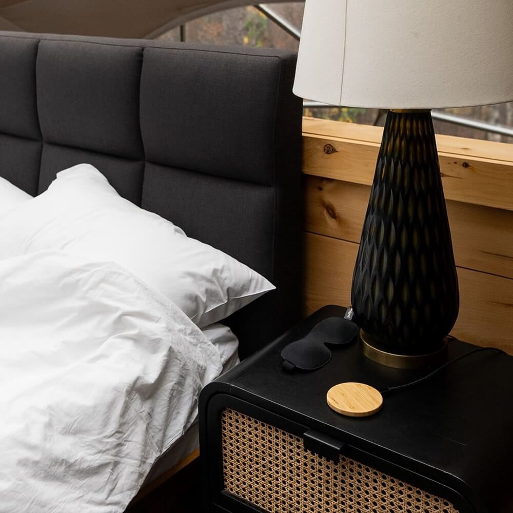 A bedside table with a black lamp, an eyes sleeping mask, and a wireless charging pad made from wood. The table is next to a bed with white linens and a gray headboard