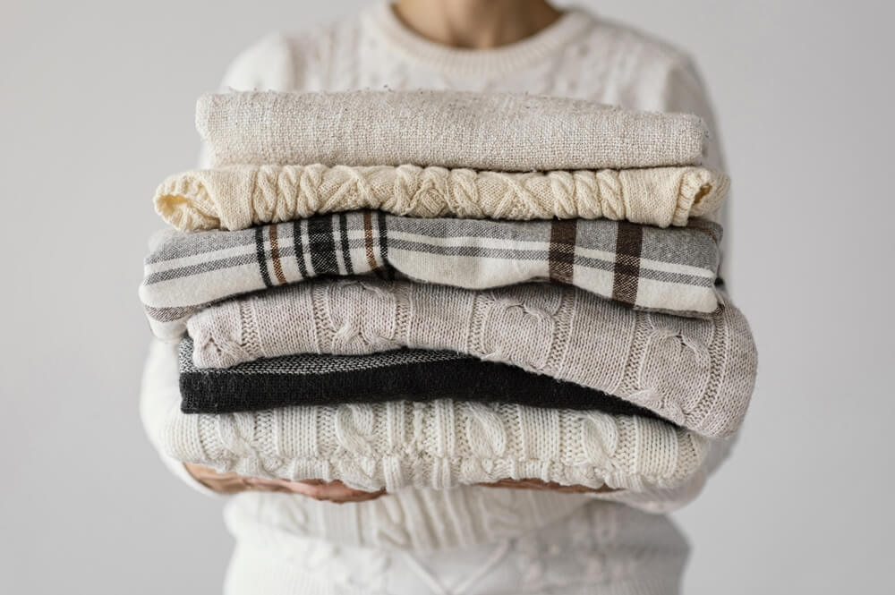 A person holding a stack of folded warm sweaters in various colors and patterns